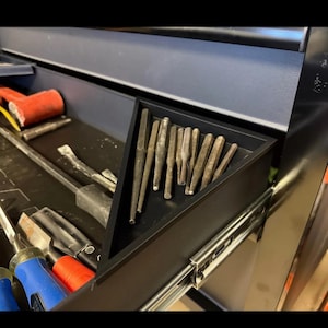 Może przedstawiać: Czarna szuflada skrzynki narzędziowej wypełniona różnymi narzędziami. Trójkątna taca zawiera kolekcję metalowych dziurkaczy. Inne narzędzia to czerwony młotek, łomy i śrubokręty z niebieskimi i czerwonymi uchwytami. Narzędzia są ułożone na czarnej powierzchni.