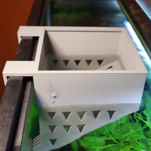 Pode incluir: Uma caixa de filtro de aquário cinza claro com recortes triangulares. O filtro é montado na lateral de um aquário de vidro, parcialmente submerso em água. Plantas aquáticas verdes são visíveis abaixo.
