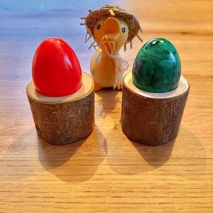 Può includere: Due uova di Pasqua, una rossa e una verde, sono esposte in porta uova di legno. Un'anatra di legno con un cappello di paglia è sullo sfondo.
