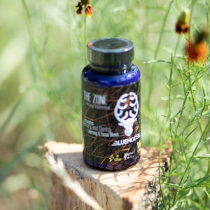 May include: A blue bottle with a black lid and a gold label that says "The Zone" and "Blue Moose". The bottle is sitting on a wooden stump in a grassy area.