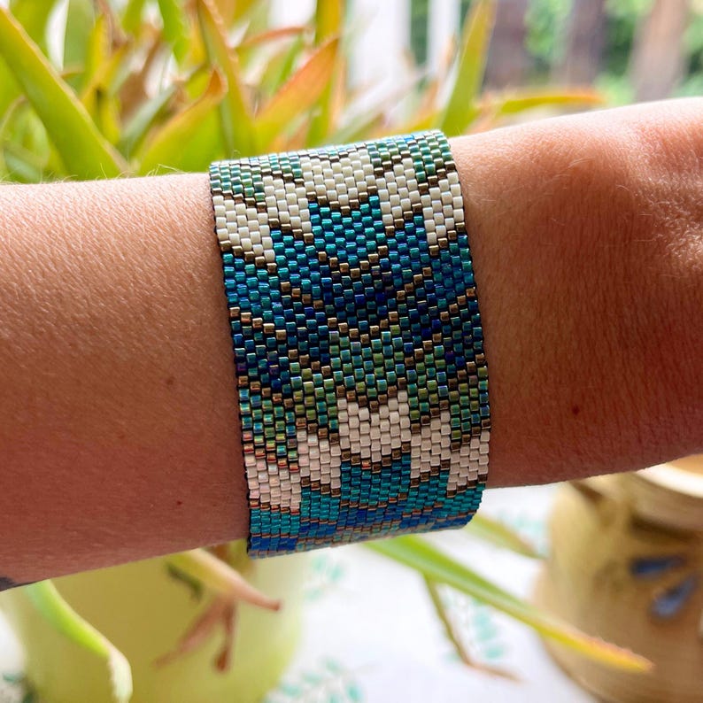 May include: A wide beaded bracelet with a geometric pattern. The bracelet features a repeating design of white, teal, and dark blue beads. The bracelet is approximately 2 inches wide.