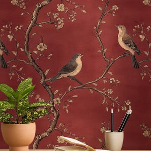 Può includere: Un ufficio domestico con una scrivania in legno, una lampada nera e un orologio bianco. Lo sfondo è una carta da parati rossa con motivi floreali e uccelli. Una camicia bianca è appesa a una scala di legno.