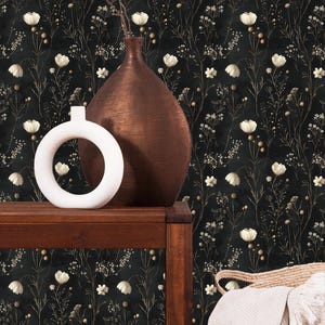 May include: A black and white floral wallpaper with a brown vase and a white ceramic ring holder on a dark wood table. A white blanket is draped over a woven basket.