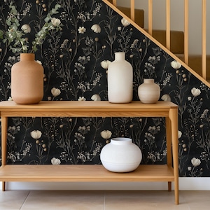 May include: A wooden console table with two shelves, featuring a black and white floral wallpaper with gold accents. The table is decorated with three ceramic vases in various shapes and sizes, two in white and one in brown.