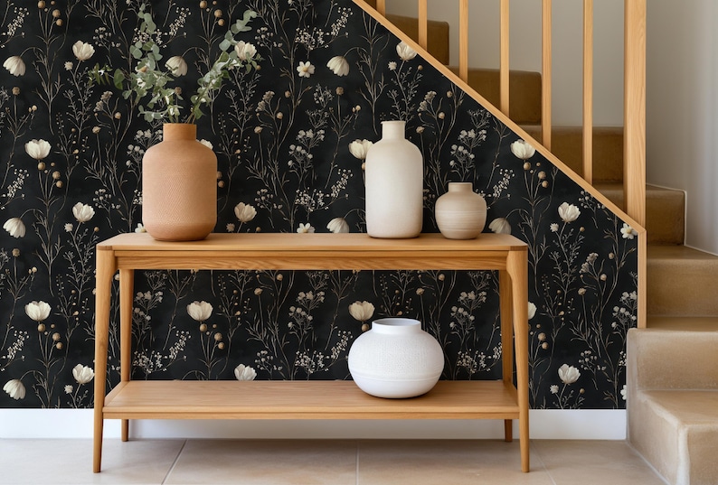 May include: A wooden console table with two shelves, featuring a black and white floral wallpaper with gold accents. The table is decorated with three ceramic vases in various shapes and sizes, two in white and one in brown.
