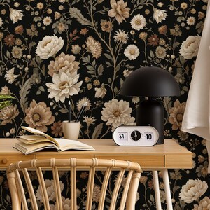May include: A home office scene featuring a wooden desk with a black lamp, a retro clock, and a potted plant. The background is a floral wallpaper with cream and brown flowers on a black background. A white shirt hangs on a wooden ladder.