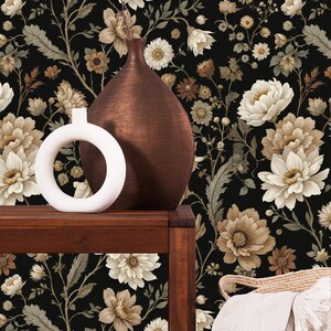 May include: A dark brown wooden table displays a white circular vase and a copper-colored vase against a black floral wallpaper background. The wallpaper features large, cream-colored flowers and smaller brown and tan blooms.