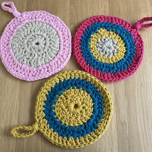May include: Three circular crochet pot holders in various colours, each with a loop handle. One is pink and beige, another is pink, teal, and yellow, and the third is yellow and teal. They rest on a light wooden surface.