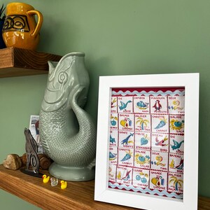 May include: A framed print with a white border displaying a vintage pattern with French words and illustrations. The print is on a wooden shelf with decorative items, including a ceramic fish sculpture and a yellow mug.