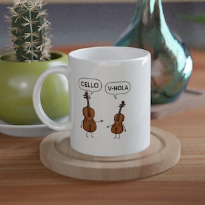 Può includere: Tazza in ceramica bianca con violoncelli dei cartoni animati e fumetti con la scritta "CELLO" e "V-HOLA". La tazza è su un sottobicchiere di legno. Un cactus e un vaso decorativo sono sullo sfondo.
