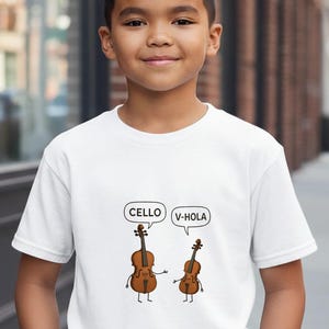 May include: White t-shirt with a cartoon cello and violin, each with speech bubbles saying "Cello" and "V-Hola". The cartoon instruments have arms and legs. The shirt is worn by a young person.
