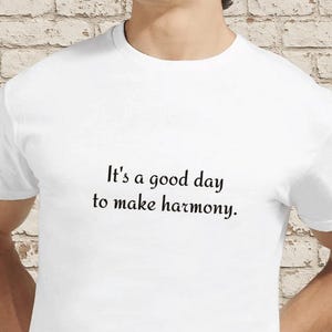 Puede incluir: Camiseta blanca con el texto "It's a good day to make harmony" en escritura negra. La camiseta es lisa y tiene cuello redondo. La imagen muestra la camiseta en una persona.