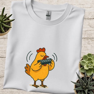 May include: White t-shirt featuring a cartoon chicken playing a harmonica. The chicken is yellow with a red comb and brown feet. The harmonica is gray. The design is centered on the shirt.