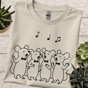 May include: Cream-colored t-shirt featuring a black line drawing of four mice wearing bow ties and singing. Above the mice are three musical notes. The shirt is surrounded by potted plants.