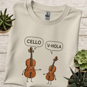 May include: Cream-coloured t-shirt with a cartoon cello and violin in speech bubbles. The cello says "CELLO" and the violin says "V-HOLA". The instruments have arms and legs, and are drawn in brown.