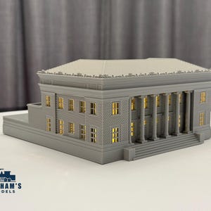 Puede incluir: Maqueta gris de un edificio clásico con columnas y ventanas. El modelo tiene un techo detallado y una pequeña estructura lateral. Las ventanas parecen iluminadas con una luz amarilla cálida. El modelo está sobre una superficie blanca.