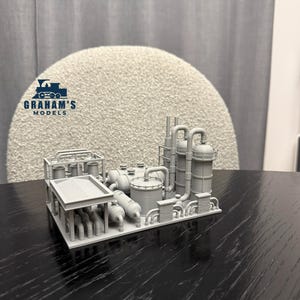 May include: A gray model of an industrial complex, featuring tanks, pipes, and towers. The model is labeled "GRAHAM'S MODELS" and sits on a dark surface. The complex includes various cylindrical and rectangular structures.