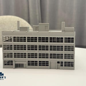 May include: A grey scale model of a multi-storey industrial factory building featuring a brick-textured facade, numerous rectangular window panes across four levels, a flat roof with chimneys, and a ground-level entrance. The logo Graham's Models is visible.