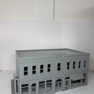 May include: A gray, two-story building model with multiple windows and a flat roof. The building features a storefront with large windows and an awning. The model is a miniature representation of a commercial structure.