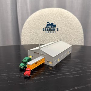 Puede incluir: Un modelo en miniatura de un almacén con techo de metal corrugado, un camión verde y un camión rojo que tira de un remolque amarillo con el nombre "Hennis" impreso. El modelo está sobre una superficie negra.