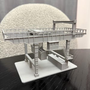 May include: A gray 3D-printed model of a gantry crane, featuring a horizontal beam supported by vertical columns. The model includes a detailed platform, a small staircase, and a container hanging from the crane. The base is a light gray square.