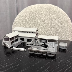 May include: A grey miniature sawmill model featuring several buildings, machinery, and stacks of timber. The model includes a building with stairs, a processing machine, and a storage area. The scene is set on a dark surface.