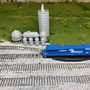 Może przedstawiać: Niebieska lokomotywa modelu pociągu Conrail z białymi paskami i numerem 1984. Model pociągu znajduje się na torze z szarym kompleksem przemysłowym w tle.