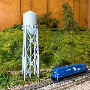 May include: A model train scene featuring a light gray water tower with a conical roof and a blue Conrail locomotive. The scene includes miniature green foliage, gravel, and train tracks, set against a wooden backdrop.
