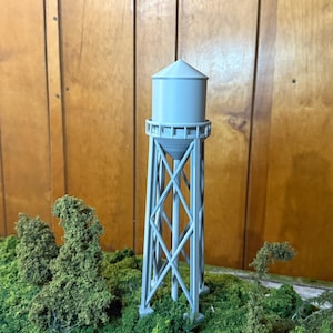 May include: A grey model water tower with a conical roof, supported by a lattice structure. The miniature tower stands on a green, textured base with small, leafy trees. The background is a wooden panel.