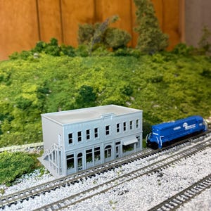 May include: A model train scene featuring a two-story gray building with arched windows and a blue locomotive. The scene includes miniature train tracks, green foliage, and a wooden backdrop, creating a detailed diorama.