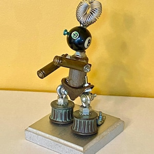 May include: A whimsical, handmade robot sculpture crafted from repurposed metal parts. The robot features a black eight-ball head, cylindrical arms, and a spring coil for hair. It stands on a rectangular base, showcasing a unique industrial aesthetic.