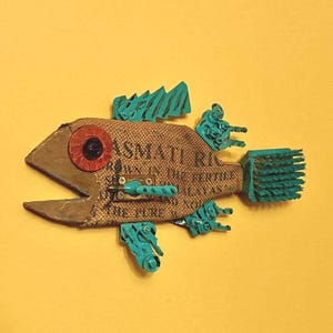 May include: A handcrafted fish sculpture made from repurposed materials. The body is constructed from a burlap sack with text, accented with brown, teal, and red elements. The fish has an open mouth and is mounted on a yellow background.