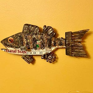May include: A handcrafted metal fish sculpture mounted on a yellow wall. The fish is constructed from various metal components, including gears, bolts, and a ruler. The sculpture features a detailed eye and a textured tail, with the text "Natural State" visible.