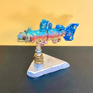 May include: A colorful, handcrafted metal sculpture of a fish. The fish is painted with blue, red, and gold hues and mounted on a silver triangular base. The sculpture has a steampunk aesthetic and is displayed against a yellow background.
