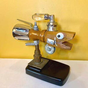 May include: A whimsical sculpture crafted from wood and metal components, resembling a stylized fish. It features a chrome faucet handle, a clear crystal-like knob, and a wooden head with a painted eye. The sculpture is mounted on a black base.