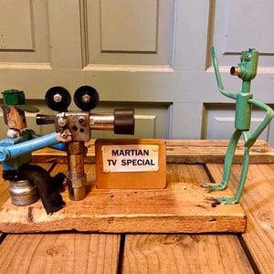 May include: A whimsical sculpture depicting a Martian TV Special. It features a camera operator, a camera, and a green alien figure. The sign reads "Martian TV Special". The figures are mounted on a wooden base.