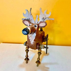 May include: A decorative deer figurine constructed from mixed materials. The body is brown with white spots, featuring a dark nose, brass-colored metal legs, and antlers fashioned from silver-toned metal keys and small wrenches attached to the head.