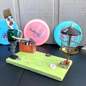 May include: A whimsical sculpture of a disc golf scene. A figure made from a paintbrush and hardware holds a toolbox of discs. A miniature disc golf basket and several discs are on a green wooden base.