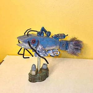 May include: A whimsical, handmade sculpture of a fish with a blue body, red eye, and metallic accents. The fish features a brush tail and is mounted on a decorative base. The sculpture has a steampunk aesthetic.