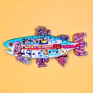May include: A decorative fish sculpture constructed from repurposed metal components and colorful collage elements. The body features the text COLLIN STREET BAKERY, a small valve handle on top, and a multi-colored tail section with various mechanical parts.
