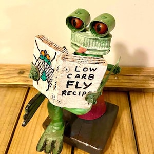 May include: A whimsical frog sculpture made from repurposed materials, holding a book titled "Low Carb Fly Recipes." The frog has green bottle cap eyes and a green body. The book features a fly illustration.