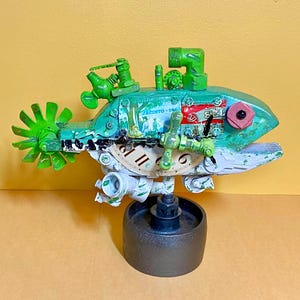 May include: A whimsical, handmade sculpture of a fish, constructed from repurposed materials. The fish features a green and white color scheme, with clock face details and various mechanical components. The sculpture is mounted on a dark gray base.