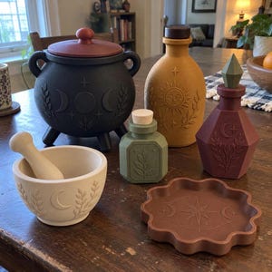 May include: A collection of decorative items including a black lidded cauldron, a mustard yellow bottle with a sun motif, a small green bottle, a maroon faceted bottle, a cream mortar and pestle, and a brown scalloped tray with a star design.