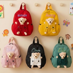 Peut inclure: Cinq sacs à dos pour enfants de couleurs vives : rouge, jaune, rose, noir et vert d'eau. Chaque sac présente un motif d'ours en peluche et un prénom brodé : Sophia, Avery, Chloe, Clara et Ella. Les ours portent des tenues en velours côtelé.