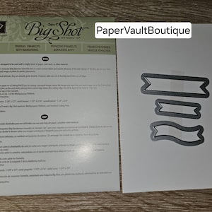 May include: A close-up of a white card with the text "PaperVaultBoutique" and a grey metal die cut of three banner shapes. The die cut is for use with a Big Shot die cutting machine.