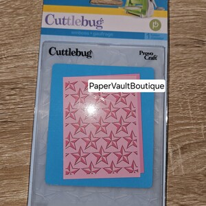 May include: A clear plastic package containing a Cufflebug embossing folder with a star pattern. The package includes a sample of the embossed design on pink paper. The text "PaperVaultBoutique" is printed on the sample.