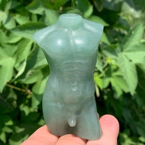 Peut inclure: Une sculpture de torse en pierre précieuse vert clair sculptée. La figurine est une représentation stylisée d'une forme masculine, avec une musculature définie. La sculpture est tenue dans une main sur fond de feuillage vert.