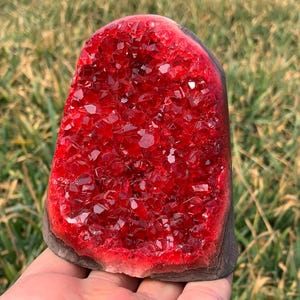 May include: A large, red crystal cluster with a rough, natural exterior. The crystal is composed of many small, faceted crystals that are tightly packed together. The crystal is a deep red colour and has a glossy finish.