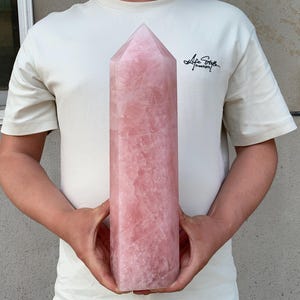 May include: A large, polished rose quartz obelisk, held by a person. The crystal is a soft pink color with visible natural veining. The obelisk is rectangular with a pointed top. The person is wearing a white t-shirt with the text "Life Style" on the left side.