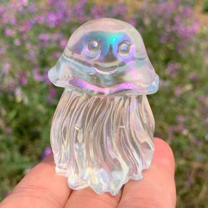 May include: Iridescent crystal jellyfish figurine with a smiling face and flowing tentacles. The clear crystal reflects a rainbow of colors, with a hint of pink and blue. The figurine is held in a hand against a blurred floral background.